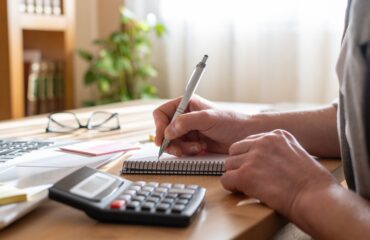 Person’s,Hands,Using,A,Calculator,And,Writing,Notes,In,A